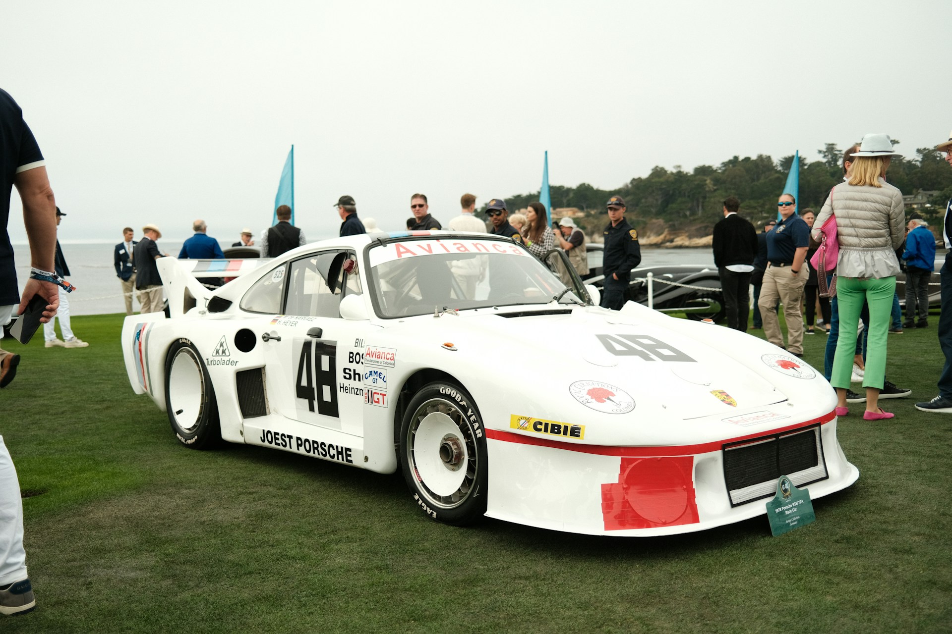 Collectors gathered around a white Porsche race car at a seaside concours